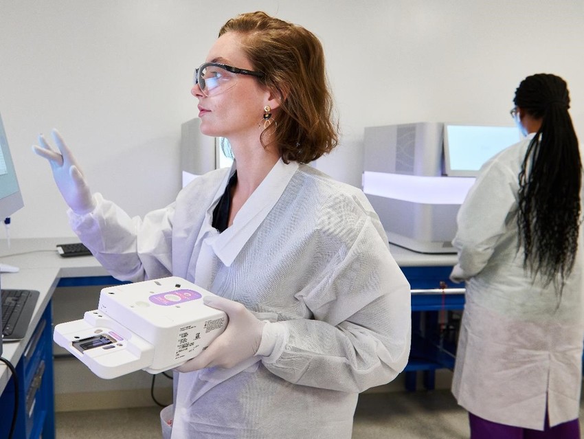A scientist in a lab coat and glove using technology in a lab setting.
