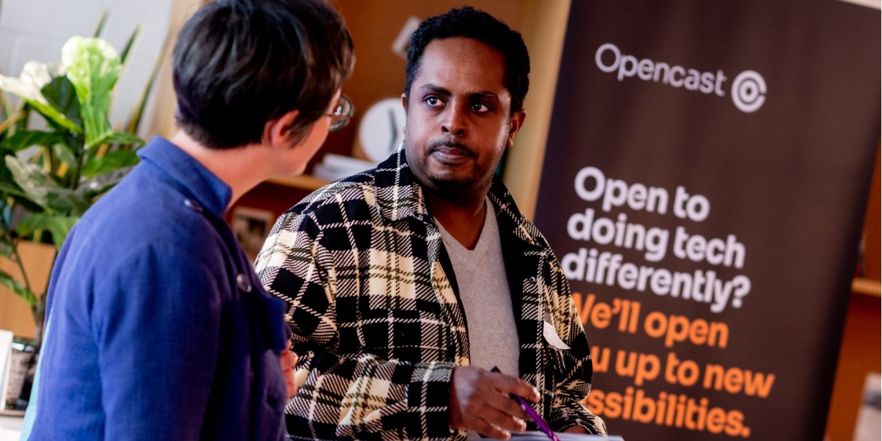 Two employees of Opencast in conversation in front of a banner that says: Opencast. Open to doing tech differently? We'll open up new possibilities.