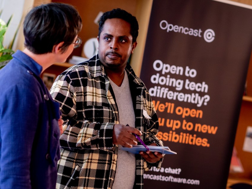 Two employees of Opencast in conversation in front of a banner that says: Opencast. Open to doing tech differently? We'll open up new possibilities.