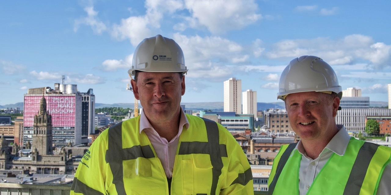 Graeme Bone, Group Managing Director, Drum Property Group (left), Dan Batterton, Head of Residential at Legal & General Investment Management Real Assets (right) in hi-vis and hard hats. They stand on top of a building with the Glasgow skyline in the background.