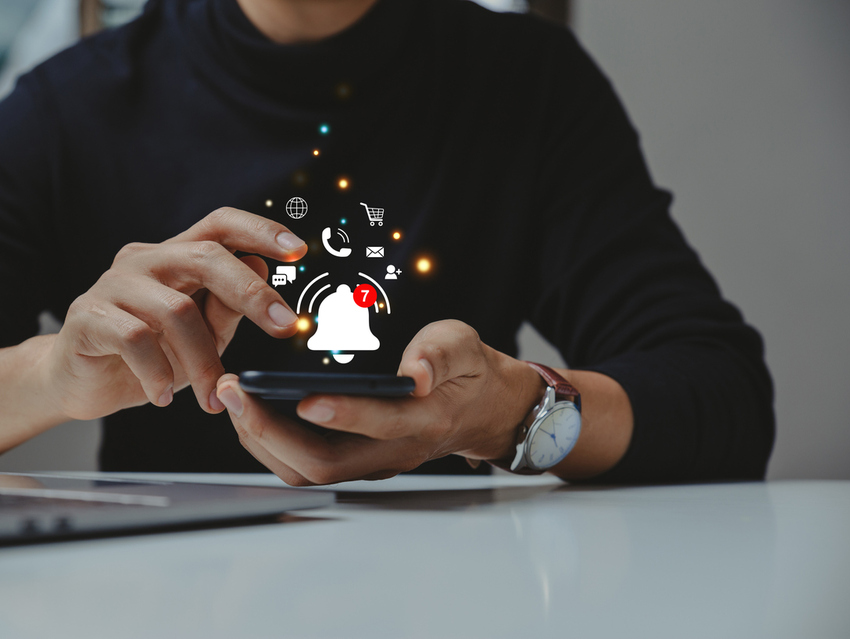 A person holding a phone in two hands in front of an open laptop. A number of alerts and messages are illustrated as incoming to the devices.