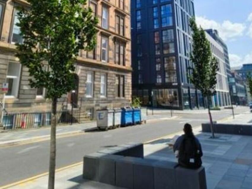 A view of the Holland Street Avenue with two young trees and a modern glass fronted building. A woman sits on new seating.