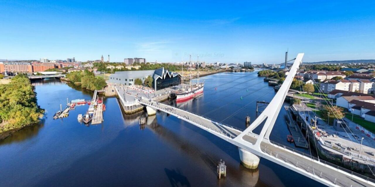 The Govan - Partick Bridge: a white bridge with a tall mast crossed the River Clyde. The Riverside Museum is on the north bank and the SEC and Science Centre can be seen in the distance. Housing lines both sides of the river.
