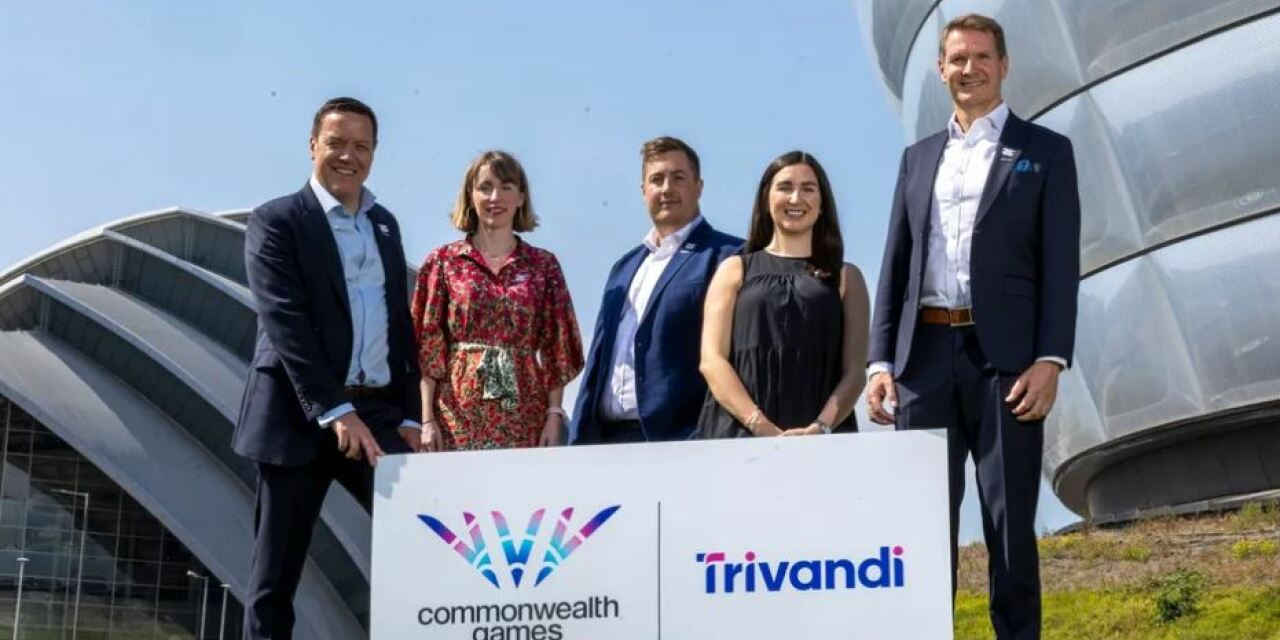 A group of officers (2 women and 3 men) from Glasgow 2026 and Trivandi stand together on the SEC campus: both the Hydro and the Armadillo are just visible. There is a white sign showing logos of both the Commonwealth Games Glasgow 2026 and Trivandi. It is a bright, sunny day with a pale blue sky.