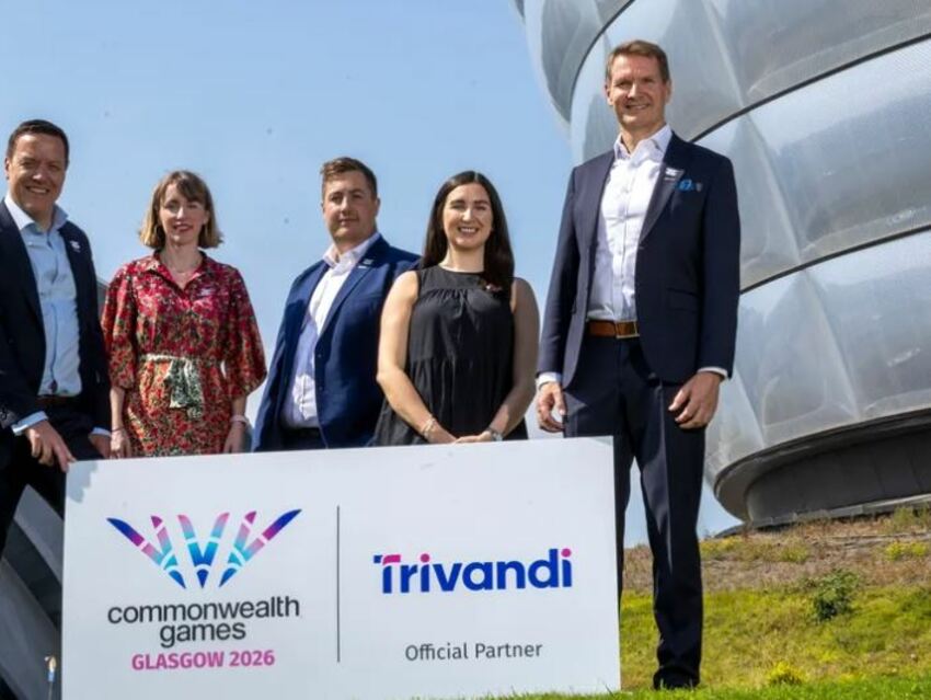 A group of officers (2 women and 3 men) from Glasgow 2026 and Trivandi stand together on the SEC campus: both the Hydro and the Armadillo are just visible. There is a white sign showing logos of both the Commonwealth Games Glasgow 2026 and Trivandi. It is a bright, sunny day with a pale blue sky.