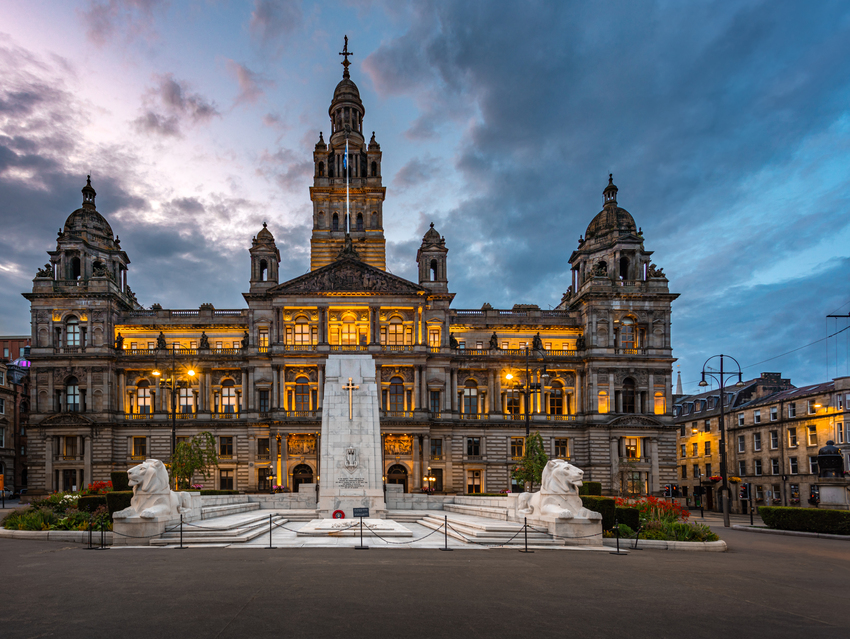 Glasgow City Chambers at dusk. The sky is darkening as night approaches with whisky clouds. The windows are illuminated yellow. The Cenotaph stands in front of the building and is white.