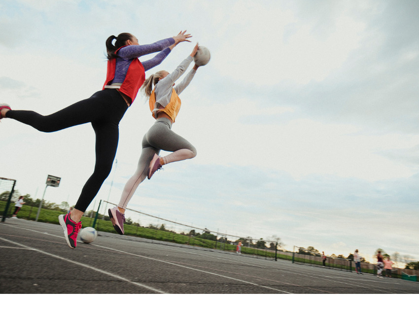 two women playing netball. they are both leaping to catch the ball against a pale blue sky.