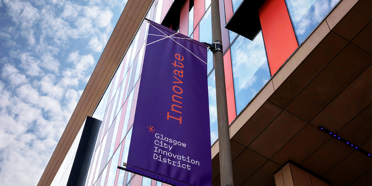A Purple Glasgow City Innovation District branded banner hangs in the street. The banner says INNOVATE and hangs in front of modern building with glass windows and orange panels (the University of Strathclyde's TIC). 
