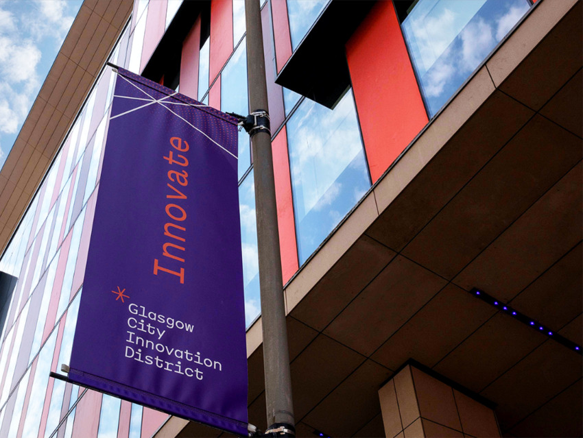 A Purple Glasgow City Innovation District branded banner hangs in the street. The banner says INNOVATE and hangs in front of modern building with glass windows and orange panels (the University of Strathclyde's TIC). 