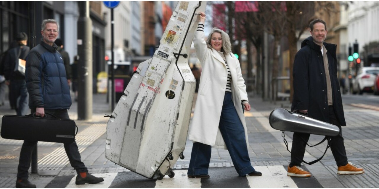 Three people carrying musical instruments walking over a crosswalk in Sauchiehall Street.