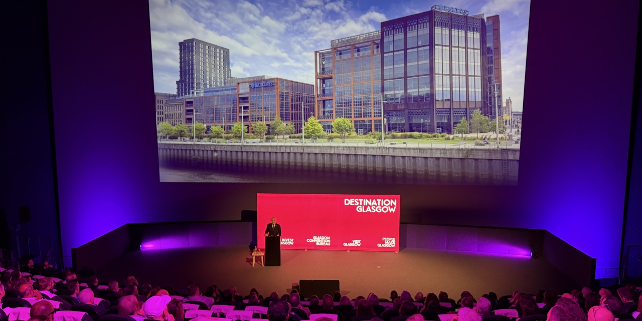 Kevin Rush, Executive Director of Economic Growth, Glasgow City Council, addressed the audience. Kevin is standing at a lectern in front of large pink screen.
